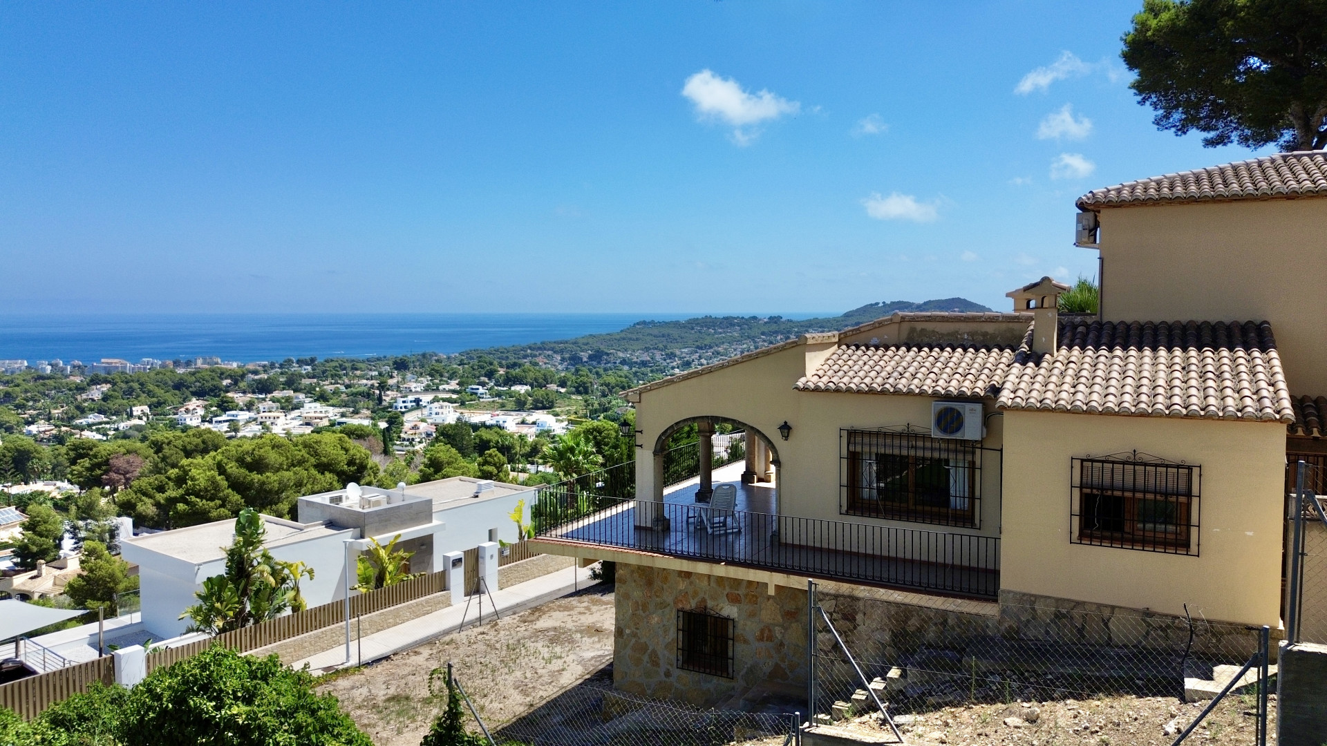 Außenansicht der Villa mit überdachter Terrasse, Bogenarchitektur und schwarzen Geländern. Das Hanggrundstück bietet Panoramablicke auf das Meer, die Stadt und die Berge.