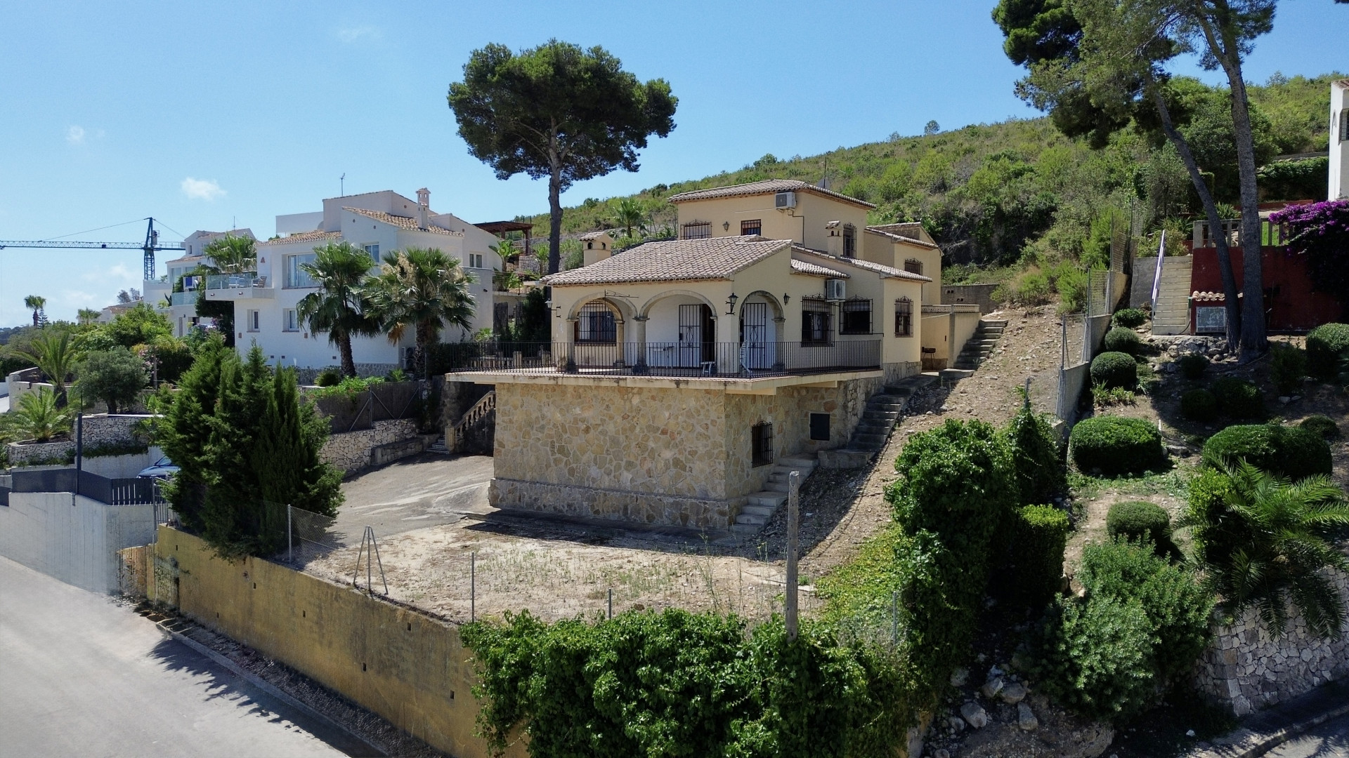 Außenansicht einer zweistöckigen mediterranen Villa mit Steinsockel und gewölbtem Balkon. Auf Hanglage mit spärlicher Vegetation unter blauem Himmel.