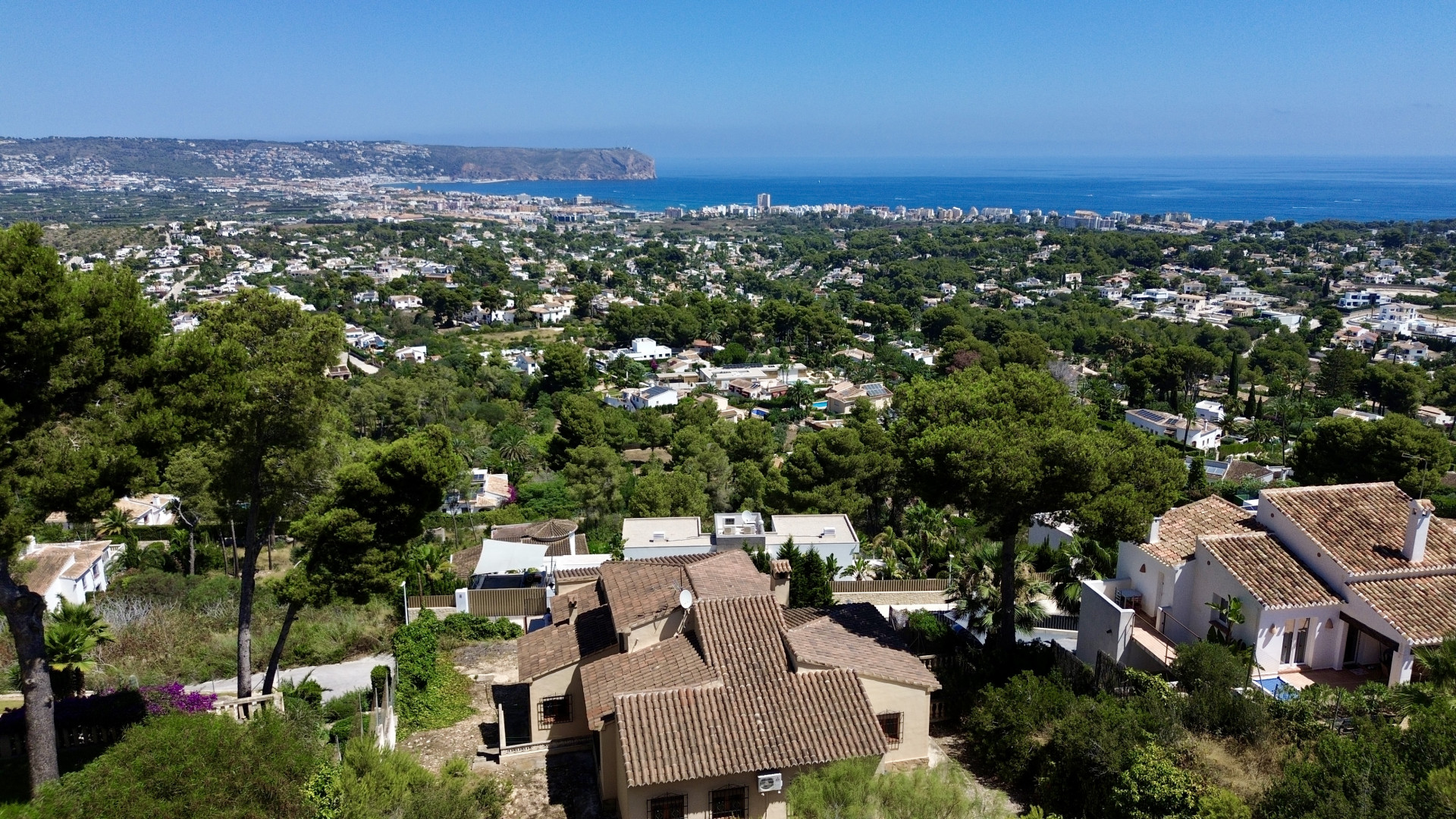 Luftaufnahme von Jávea, die traditionelle Villen mit Terrakottadächern zwischen Pinien zeigt, mit dem Mittelmeer und dem Cabo de San Antonio im Hintergrund.