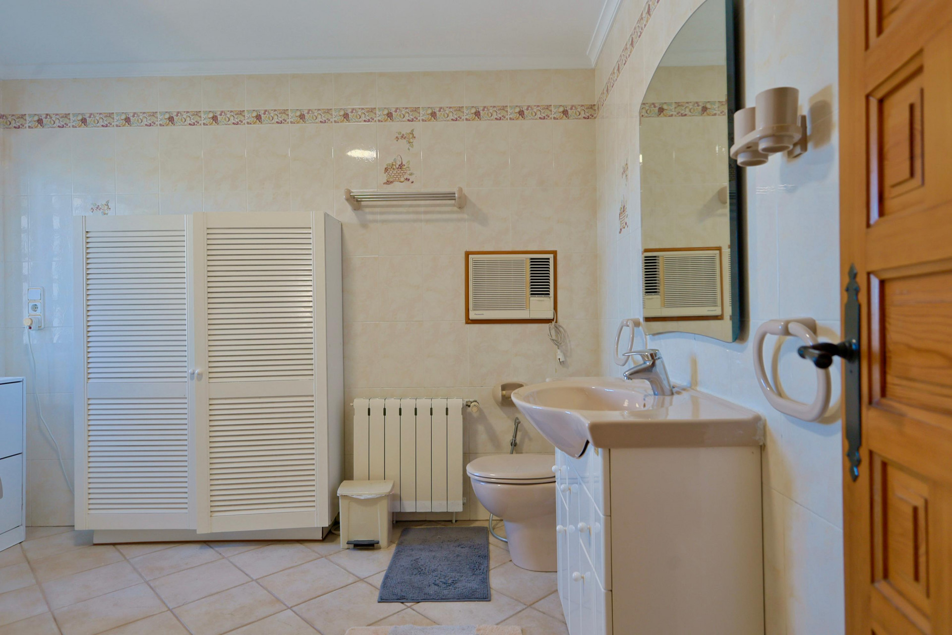 Gefliestes Badezimmer mit weißem Lamellenschrank und beigem Waschtisch.