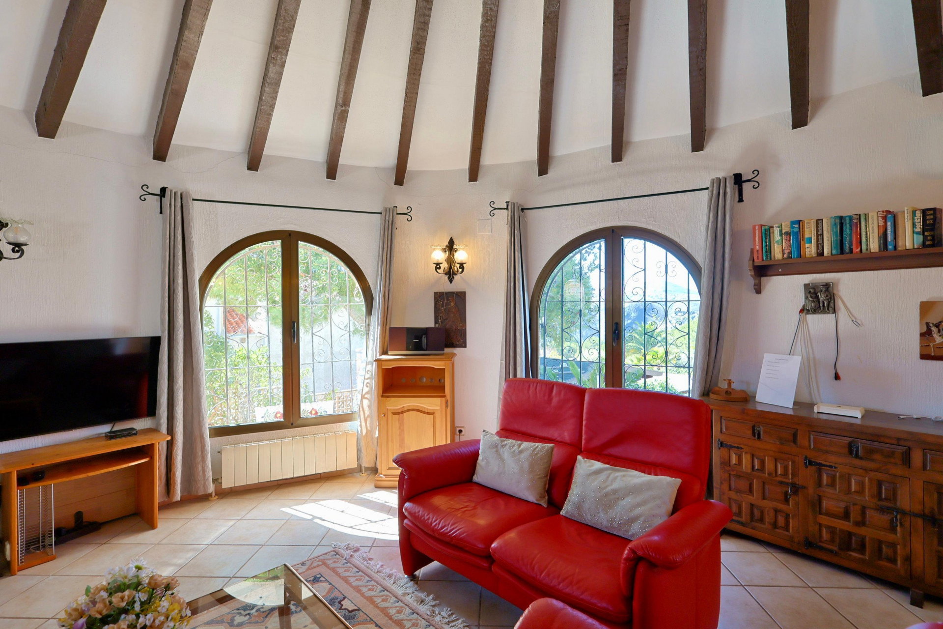 Helles Wohnzimmer mit Holzbalken, rotem Ledersofa und Bogenfenstern mit Blick auf den Garten.