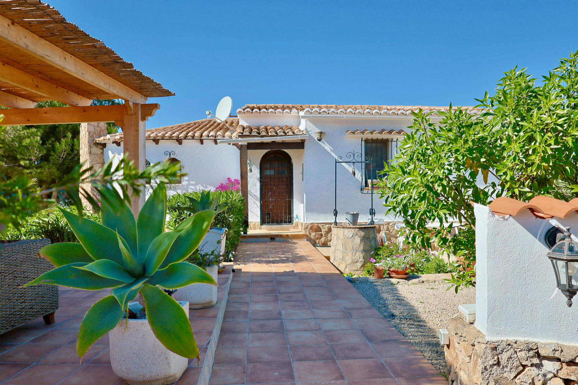 Villeneingang mit gefliestem Weg, mediterranem Grün und Steinbrunnen.