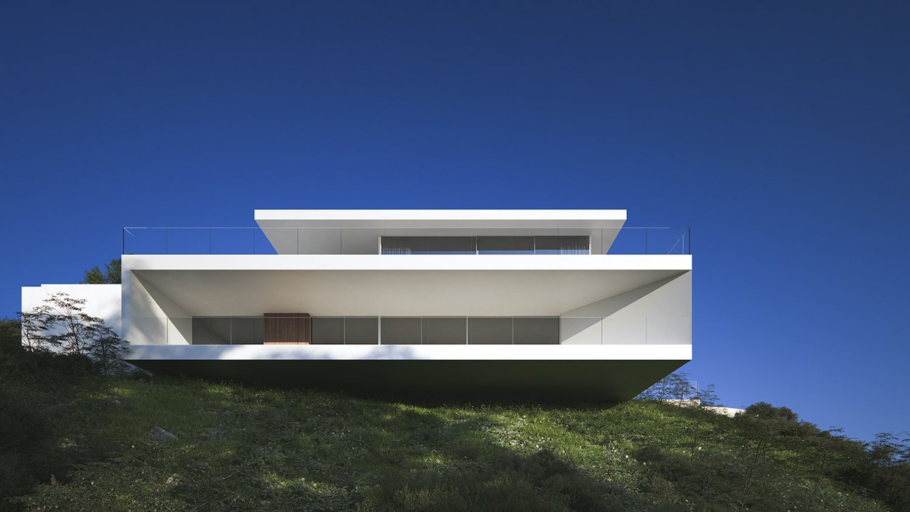 Außenfassade im minimalistischen Design mit weißen geometrischen Auskragungen und Glasgeländern, eingebettet in einen natürlichen Hang.