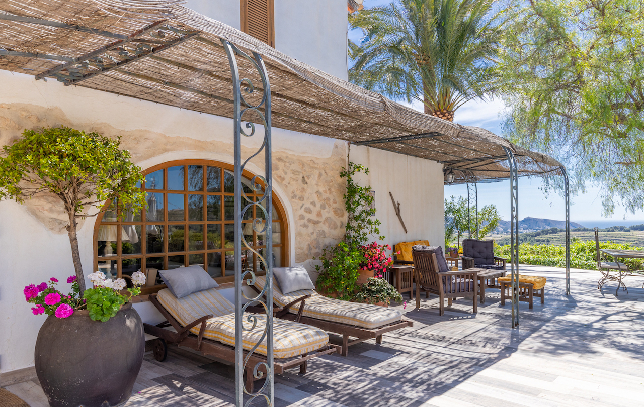 Außen-Terrasse mit einer Pergola aus Schilfrohr und Schmiedeeisen, Sonnenliegen vor einem Rundbogenfenster und Panoramablick auf das Tal und das Meer.