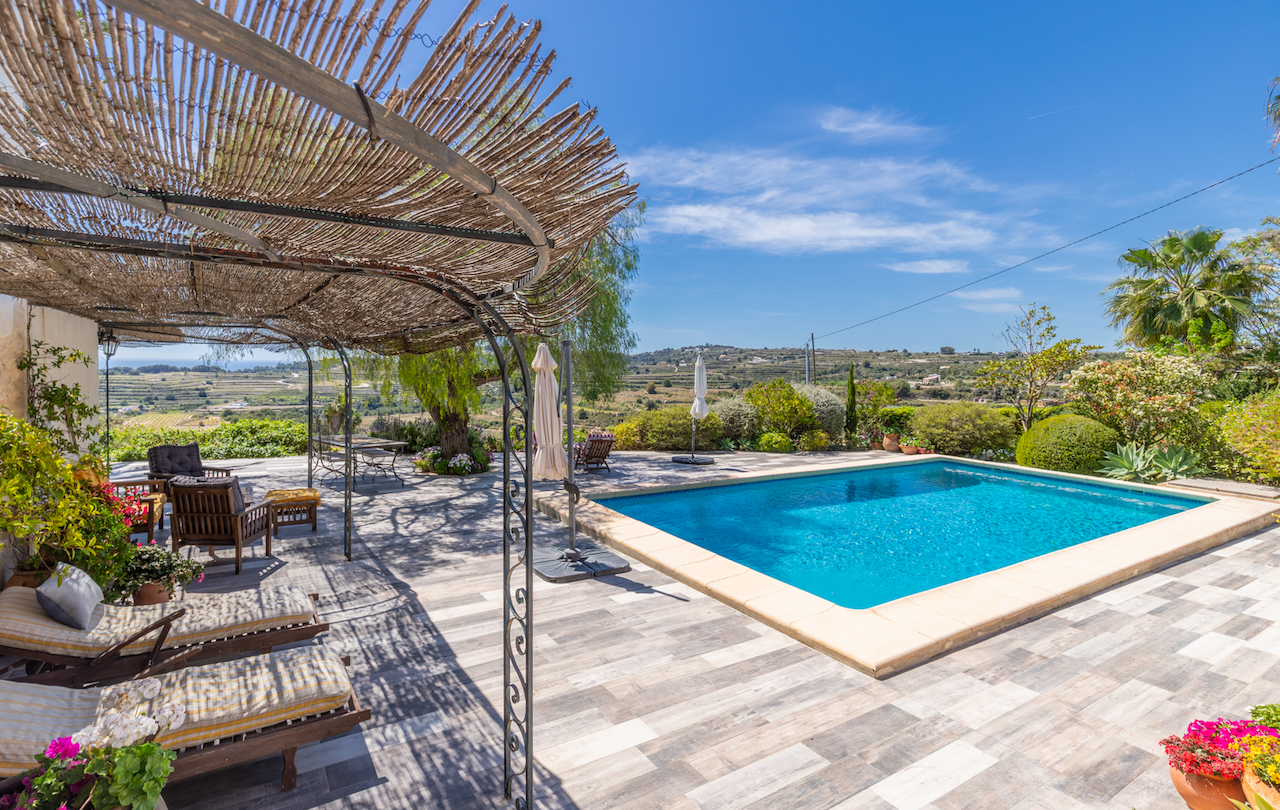 Außenbereich mit Pool und einer Pergola aus Schmiedeeisen mit Schilfrohrdach. Die rutschfeste Terrasse bietet einen weiten Blick über das Tal und die mediterrane Gartenanlage.