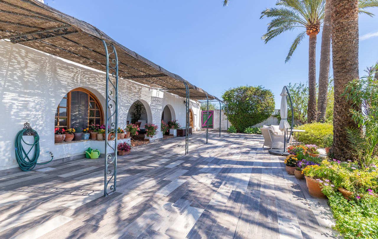 Außenterrasse mit Bodenfliesen in Holzoptik, Schilfrohr-Pergola auf Schmiedeeisensäulen und weißer Fassade mit traditionellen Rundbögen und Pflanztöpfen.