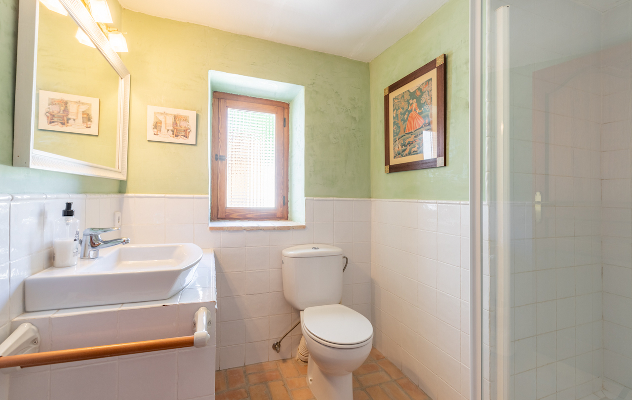 Badezimmer im rustikalen Stil mit hellgrünen Wänden, weißen Fliesen, Terrakottaboden und einem Holzfenster, das für natürliches Licht sorgt.
