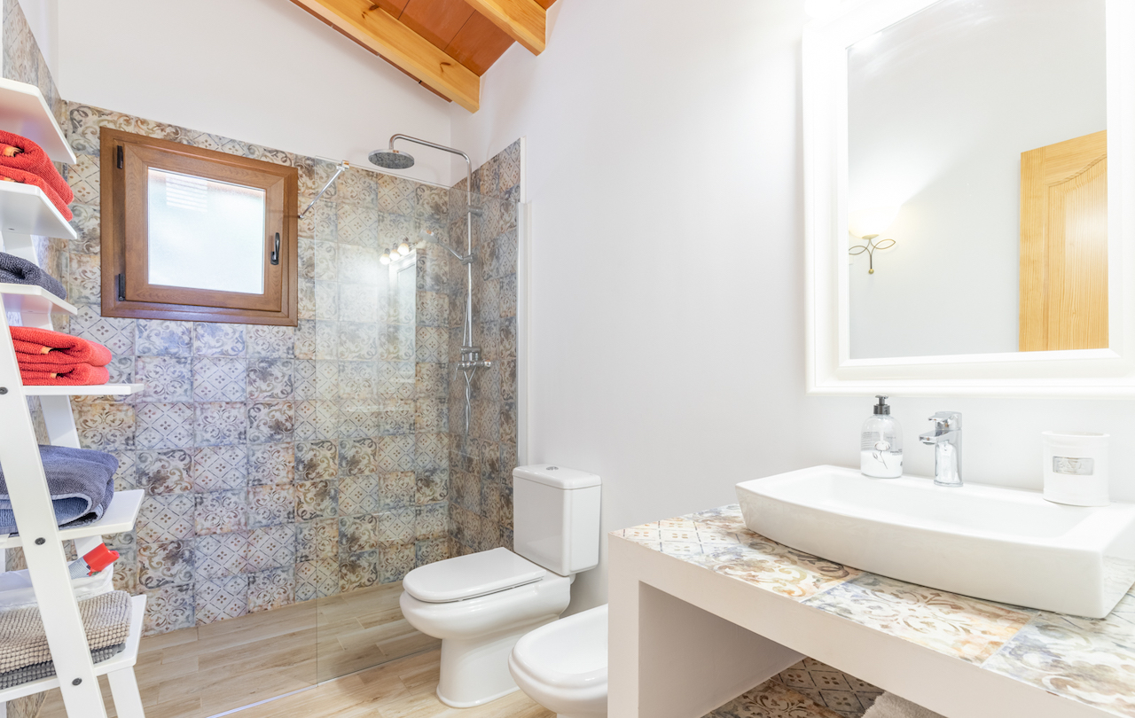 Badezimmer mit Holzbalkendecke, begehbarer Dusche mit gemusterten Fliesen und passendem Fliesentisch mit Aufsatzwaschbecken und weißen Sanitärobjekten.