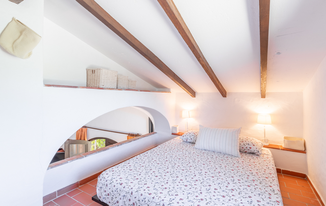 Dachgeschoss-Schlafzimmer mit sichtbaren Holzbalken, Terrakottaboden und einem markanten Rundbogen mit Blick in das darunter liegende Stockwerk.