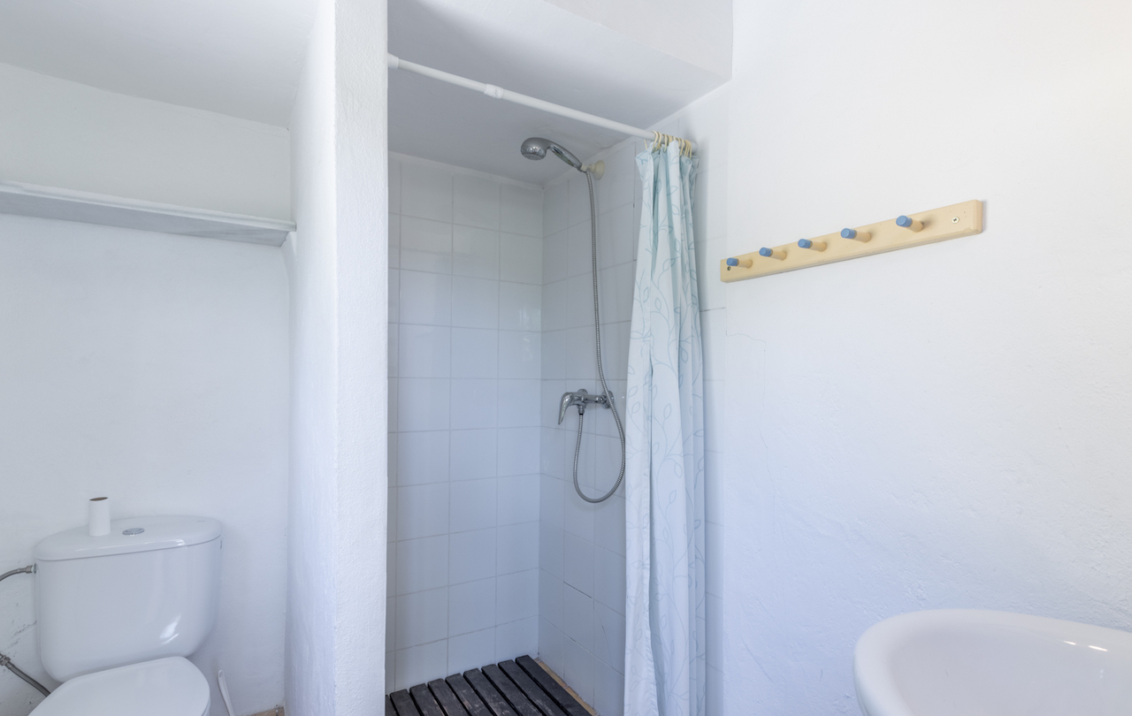 Funktionales Badezimmer mit gefliester Dusche, dunklem Holzbodenrost, gemustertem Vorhang und einer Wandgarderobe aus Holz mit blauen Haken.