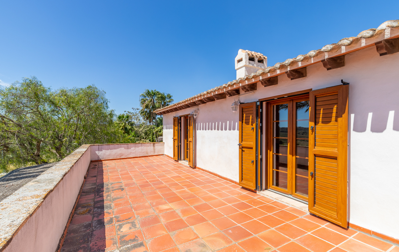 Obere Terrasse mit Terrakotta-Fliesen, sichtbaren Holzbalken unter dem Dach und traditionellen Holz-Fenstertüren mit Fensterläden vor weißer Fassade.