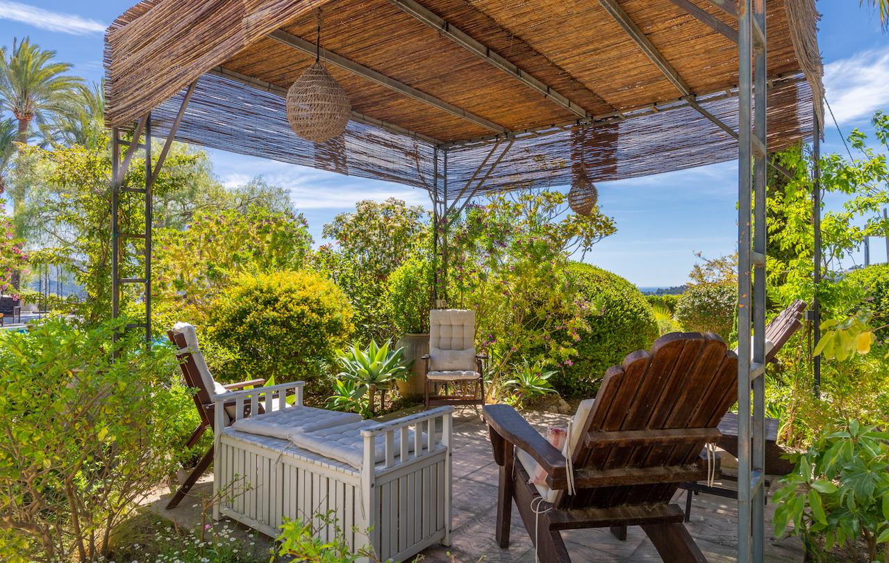 Pergola mit Metallstruktur und Schilfdach, ausgestattet mit Sitzgelegenheiten inmitten eines mediterranen Gartens mit weitem Meerblick.