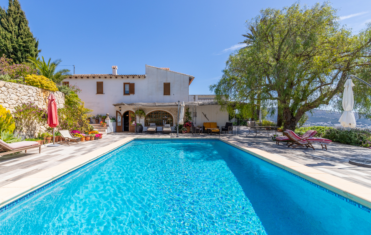Rechteckiger Swimmingpool auf einer großzügigen, rutschfesten Fliesenterrasse, angrenzend an die traditionelle weiße Fassade und einen Garten mit Natursteinmauern.