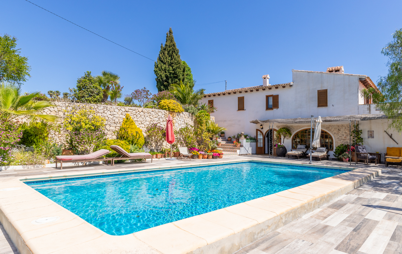 Rechteckiger Swimmingpool mit rutschfester Terrasse, Natursteinmauern und mediterraner Bepflanzung vor der traditionellen, weiß getünchten Fassade der Finca.