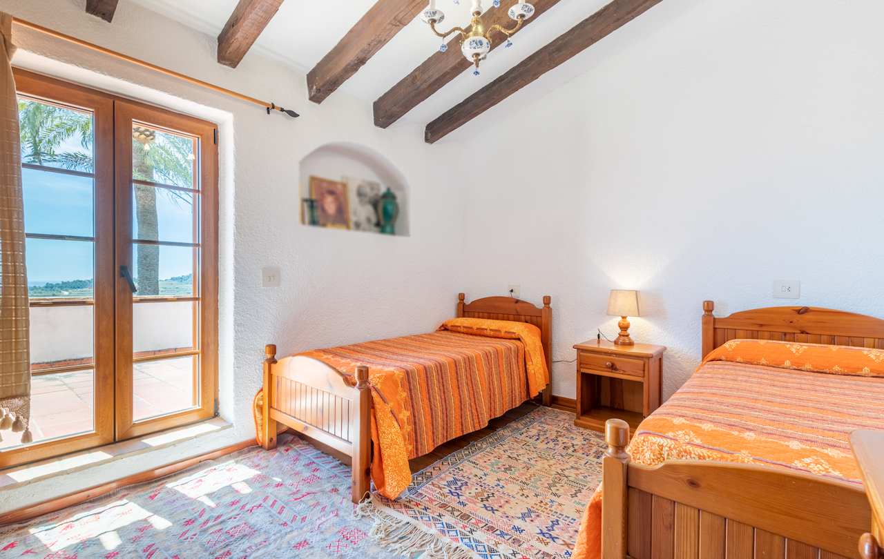 Rustikales Schlafzimmer mit Sichtbalkendecke, zwei Einzelbetten und einer Wandnische. Die Fenstertür führt direkt auf die Terrasse mit Blick auf Palmen und die Umgebung.