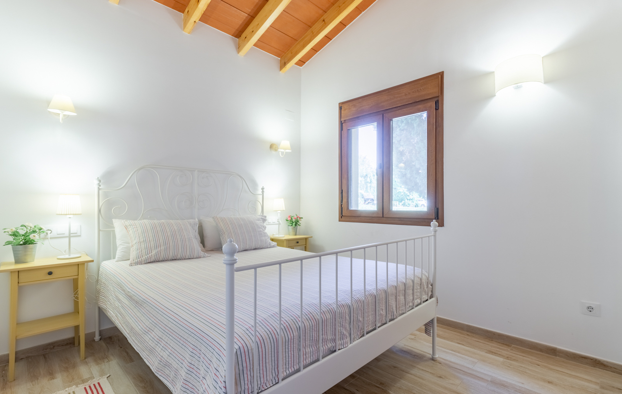 Rustikales Schlafzimmer mit sichtbaren Holzbalken, einem weißen Metallbett und Fensterrahmen aus Holz in hellem Ambiente.