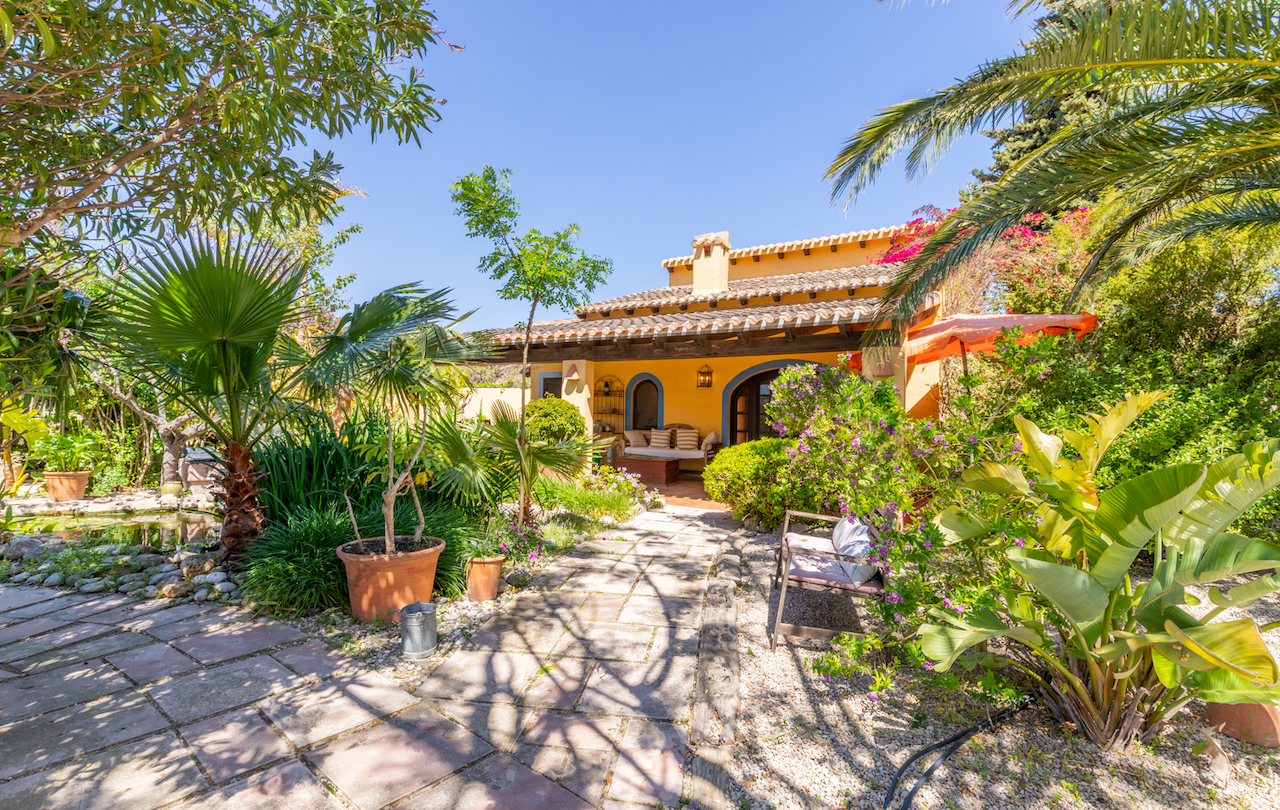 Traditionelle Fassade mit Holzbalken-Veranda in einem mediterranen Garten mit Teich, Palmen und Wegen aus Natursteinplatten.
