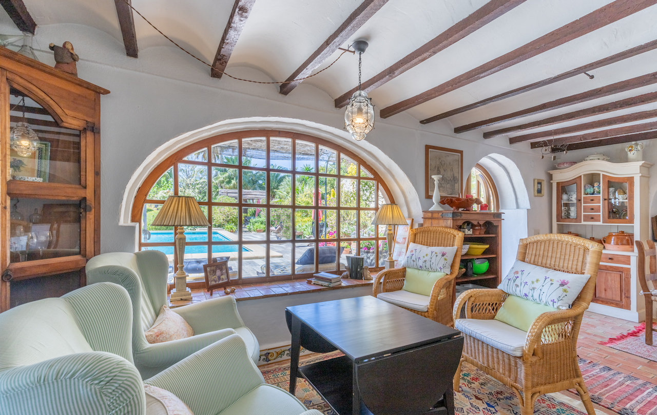 Traditioneller Wohnbereich mit sichtbaren Holzbalken, Terrakottaboden und einem großen Rundbogenfenster mit Blick auf den Pool und den mediterranen Garten.