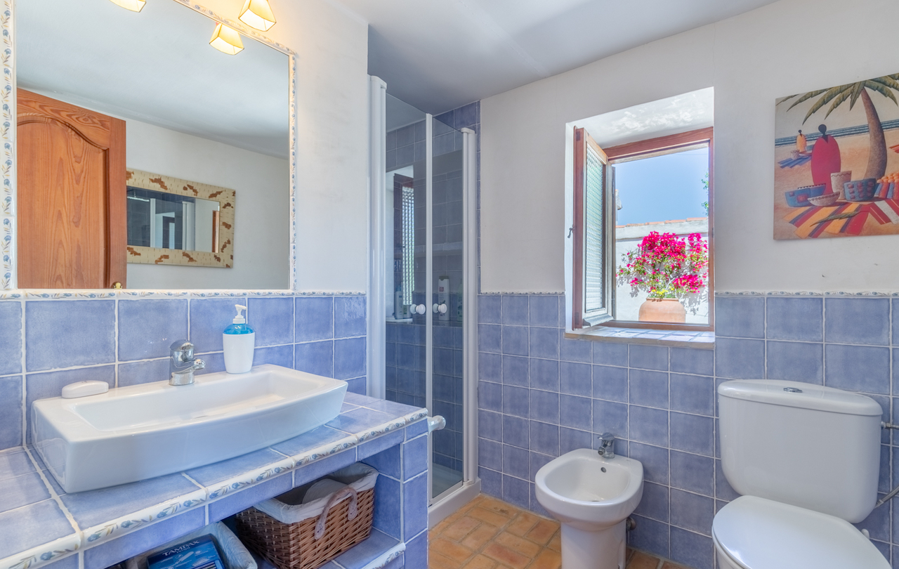 Traditionelles Badezimmer mit blauen Wandfliesen, Terrakottaboden und Holzfenster zum Innenhof. Es bietet ein Aufsatzwaschbecken, ein Bidet und eine gläserne Duschkabine.