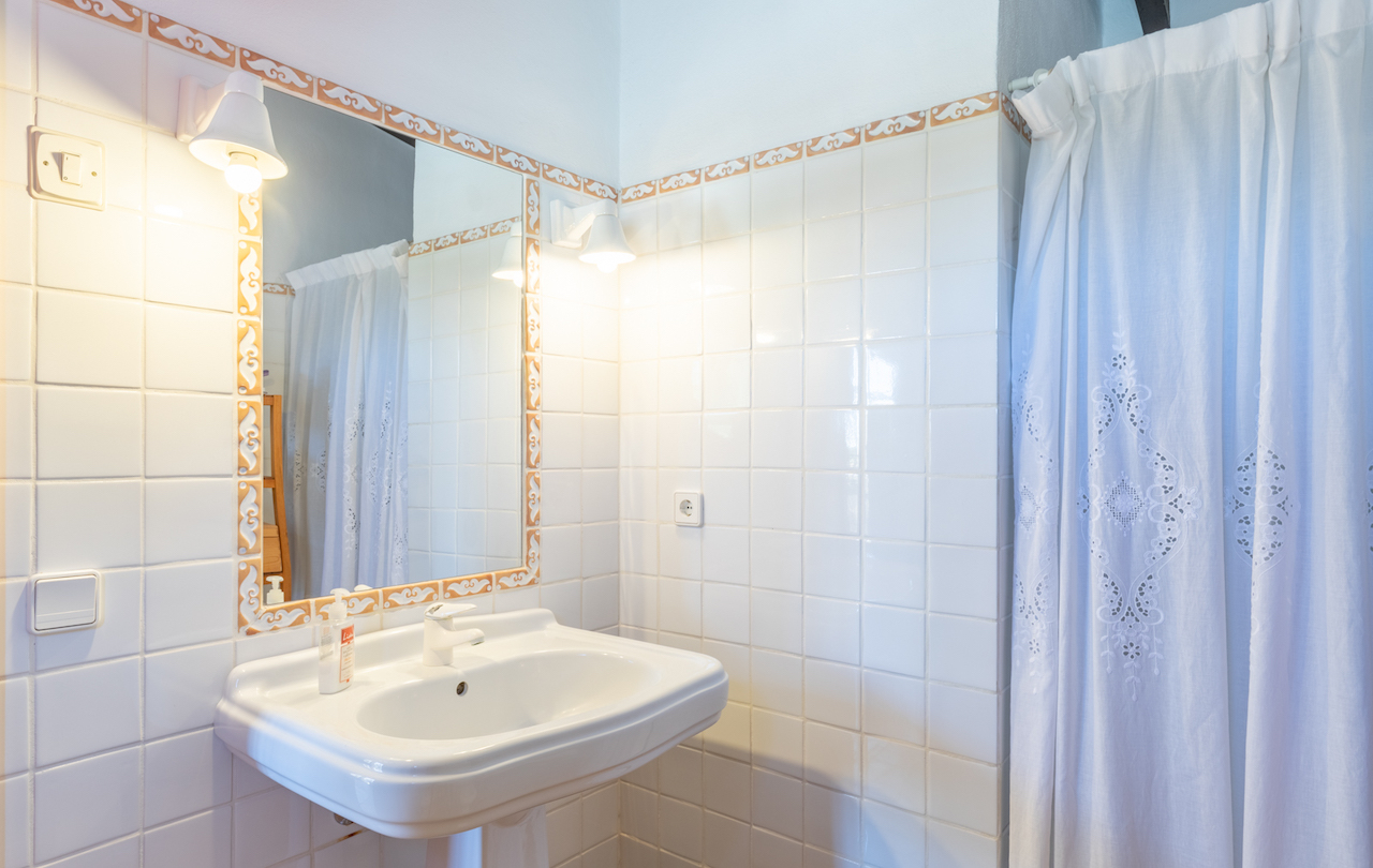Traditionelles Badezimmer mit weißem Säulenwaschbecken, dekorativ umrahmtem Spiegel und klassischen Wandleuchten neben einem bestickten Vorhang.
