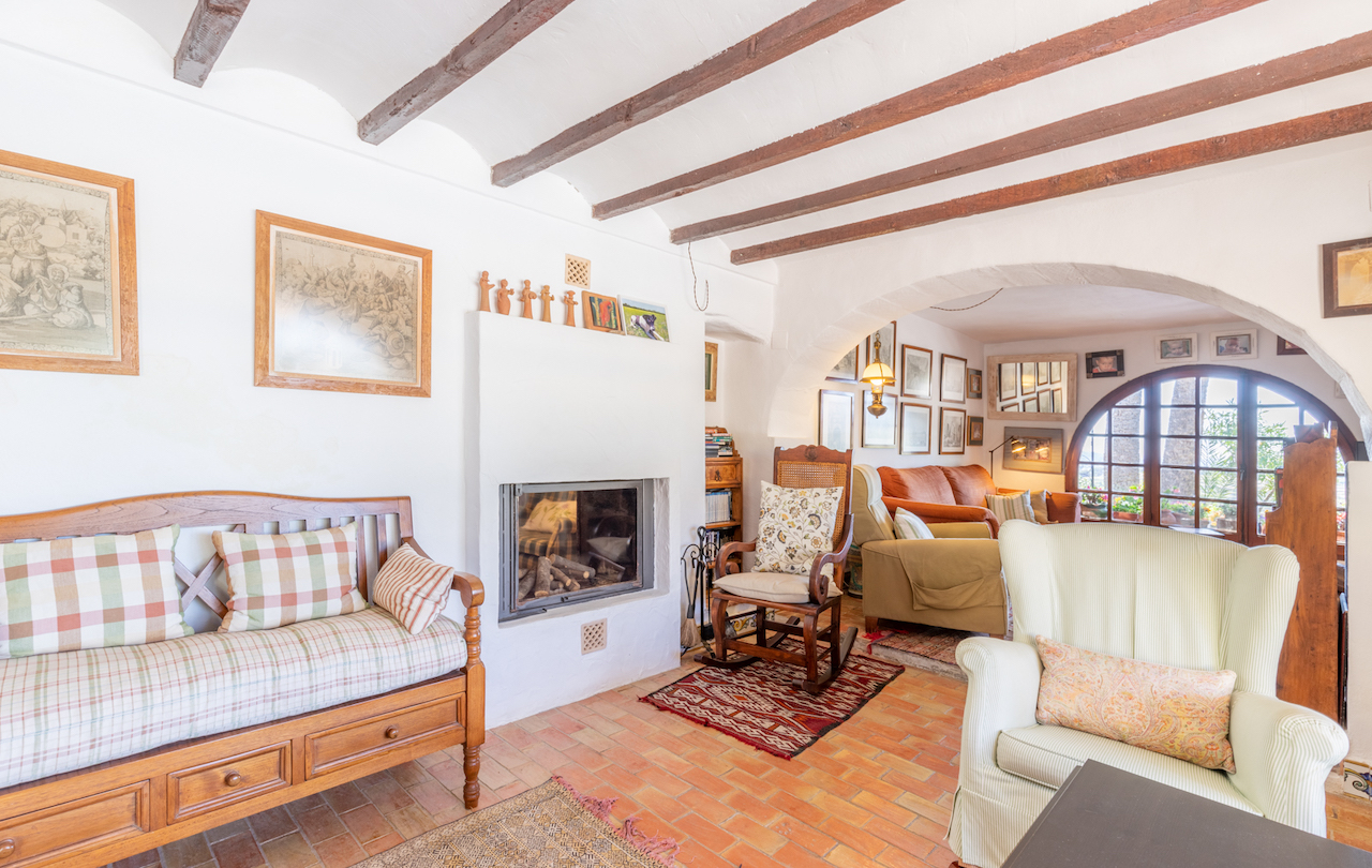 Traditionelles Wohnzimmer mit Sichtbalkendecke, Terrakottaboden und Einbaukamin. Ein Rundbogen führt zu einem hellen Sitzbereich mit großem Rundbogenfenster.