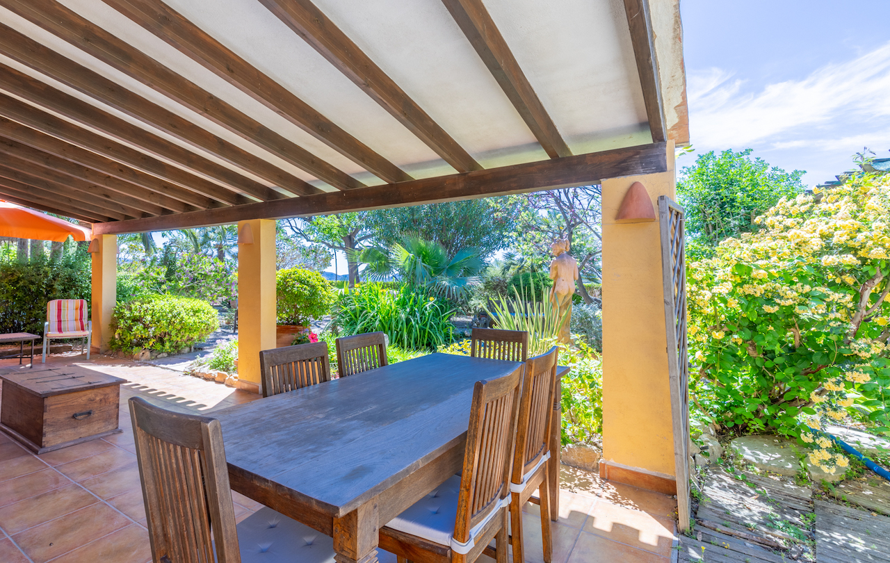 Überdachte Terrasse mit massiven Holzbalken und Terrakottaboden, ausgestattet mit einem Essbereich im Freien und Blick in den üppigen mediterranen Garten.