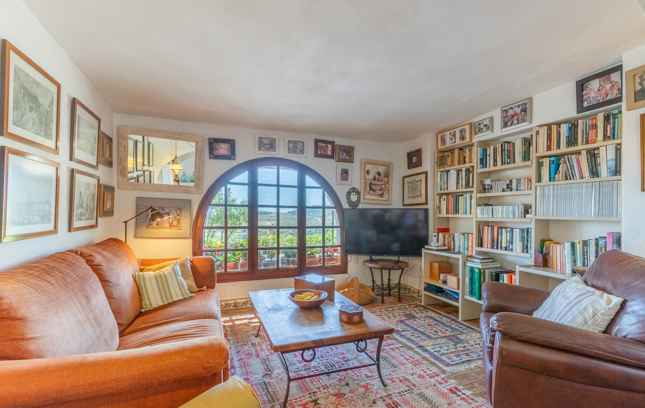 Wohnzimmer-Bibliothek mit großem Rundbogenfenster aus Holz, Einbauregalen, Terrakottaboden mit Teppichen und weitem Blick auf die umliegende Landschaft.