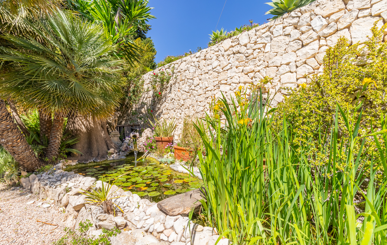 Zierteich mit Seerosen und kleinem Wasserfall, umgeben von mediterraner Vegetation und einer traditionellen Trockensteinmauer.