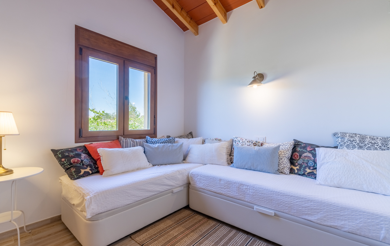 Zimmer mit L-förmigem Daybed, Decke mit sichtbaren Holzbalken und einem Holzfenster mit Blick ins Grüne. Die Einrichtung umfasst gemütliche Textilien.