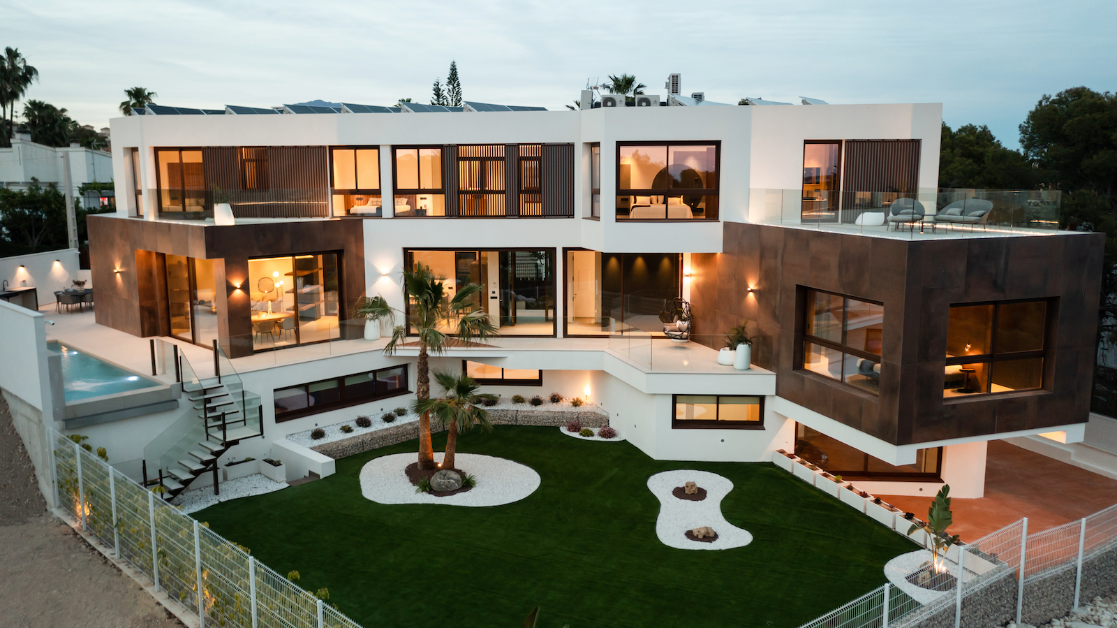 Außenansicht der modernen Villa mit mediterranem Garten, Pool und beleuchteten Terrassen.