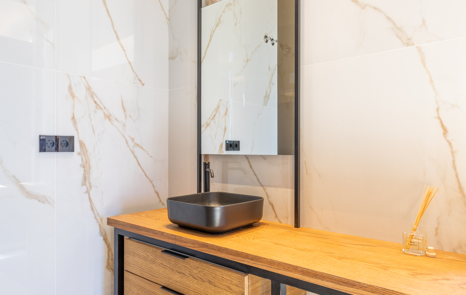 Badezimmerdetail mit mattschwarzem Waschbecken auf heller Holzablage und Wand aus geädertem Crememarmor.