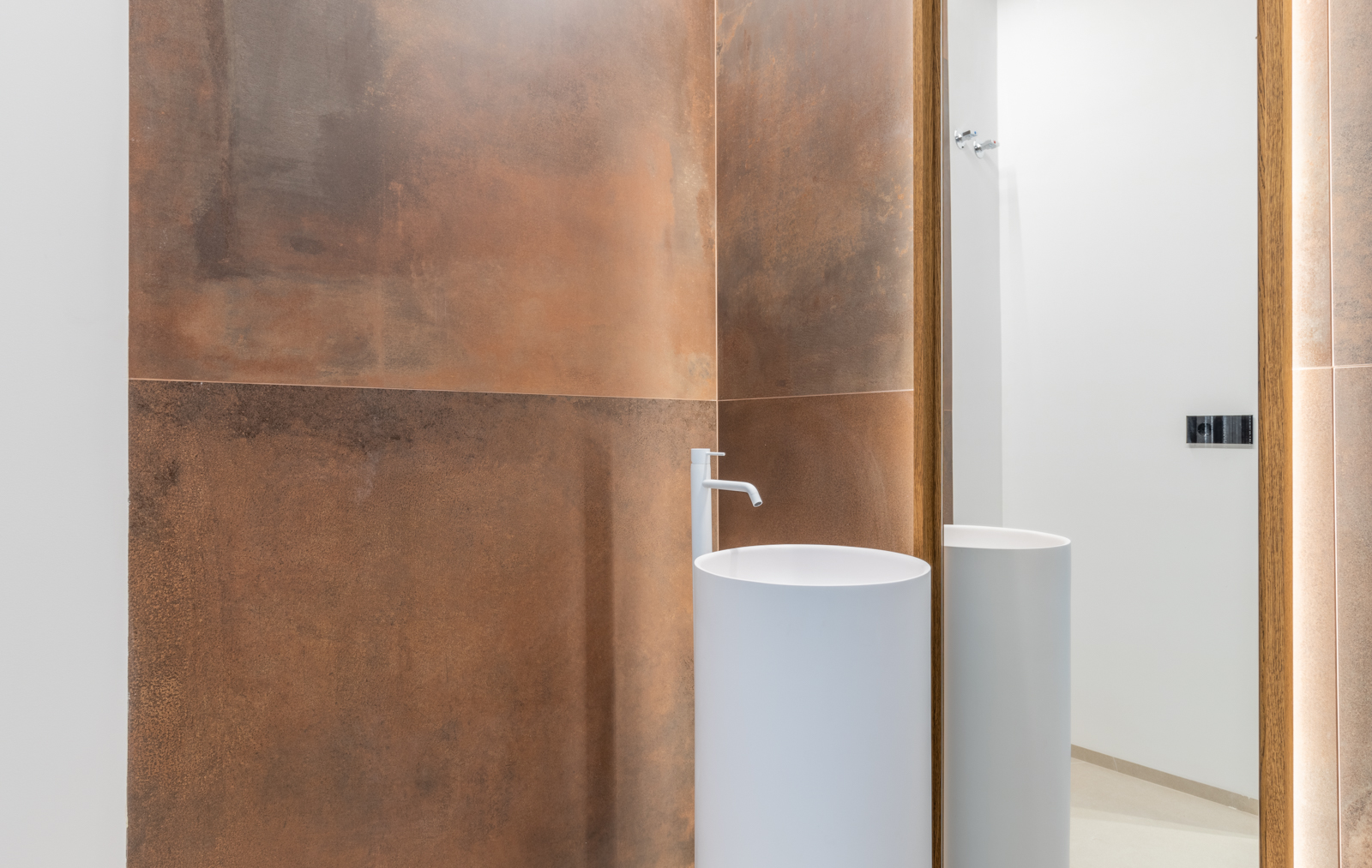 Badezimmerdetail mit weißem zylindrischem Waschbecken und rostfarbener Fliesenwand mit hinterleuchtetem Spiegel.