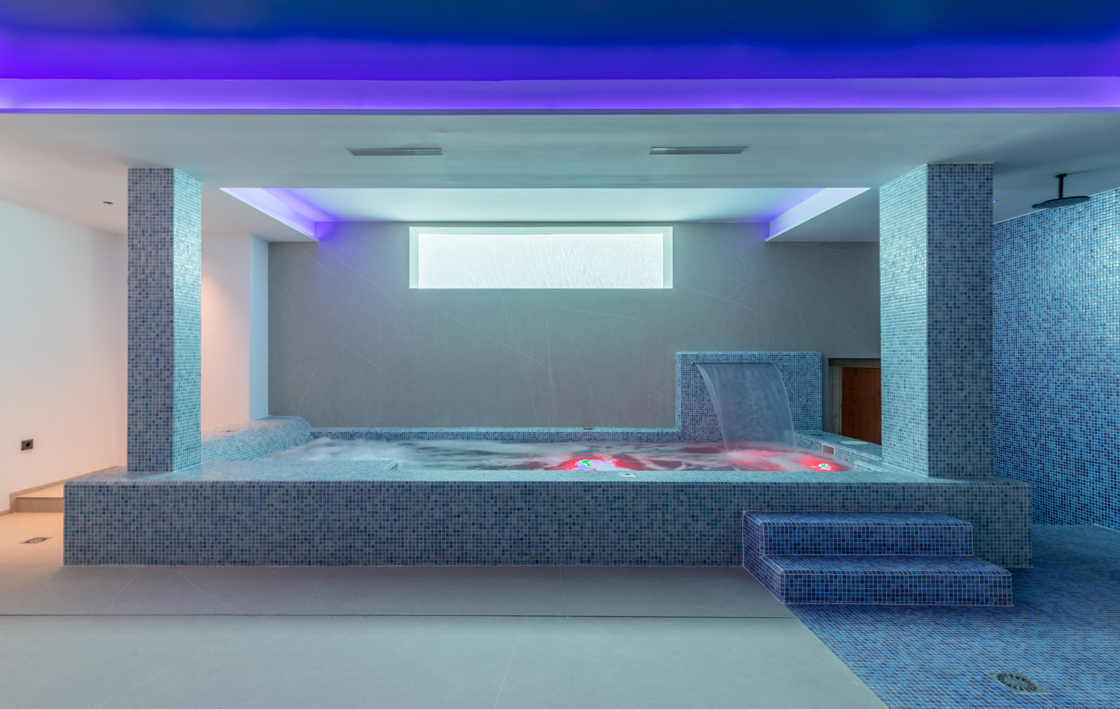Detail des Indoor-Spas mit Jacuzzi, Wasserfall und Dusche, mit blauen Mosaiken und Ambientebeleuchtung.