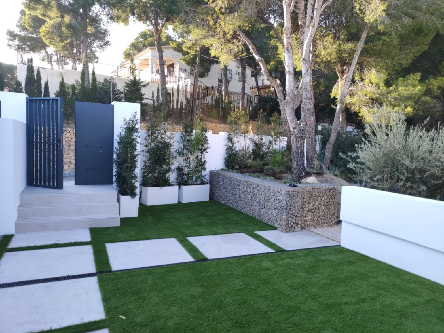 Außenbereich mit modernem Metalltor, Steinstufen und angelegtem Garten mit Kunstrasen, Betonplatten und einer dekorativen Gabionenwand unter Pinienbäumen.