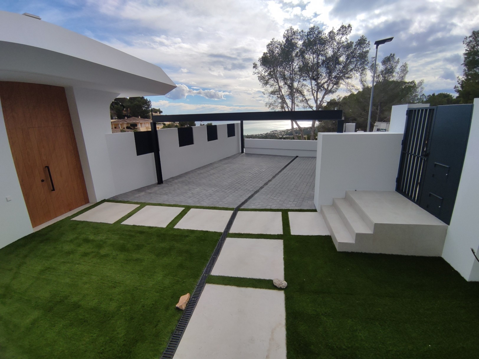 Außeneingang mit Massivholztür, gepflasterter Auffahrt und Kunstrasen mit weißen Betonplatten. Die moderne weiße Architektur gibt den Blick auf das Meer frei.