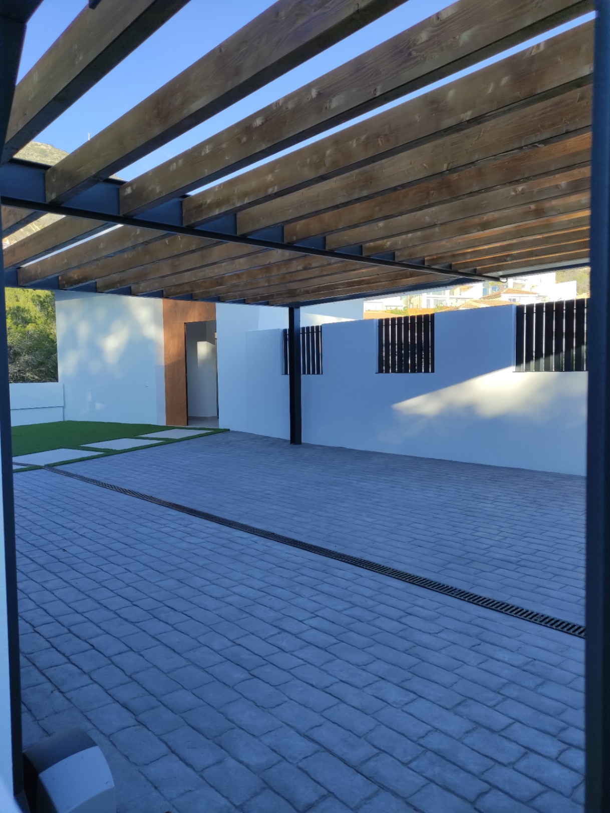 Außenparkplatz mit einer Pergola aus Holzbalken, grauem Kopfsteinpflaster und direktem Zugang zum Haus mit Akzenten aus Naturholz.