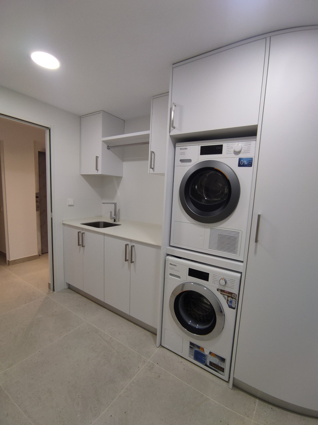 Funktionaler Hauswirtschaftsraum mit Miele-Waschsäule, minimalistischen weißen Schränken, integriertem Spülbecken und großformatigen Feinsteinzeugfliesen.
