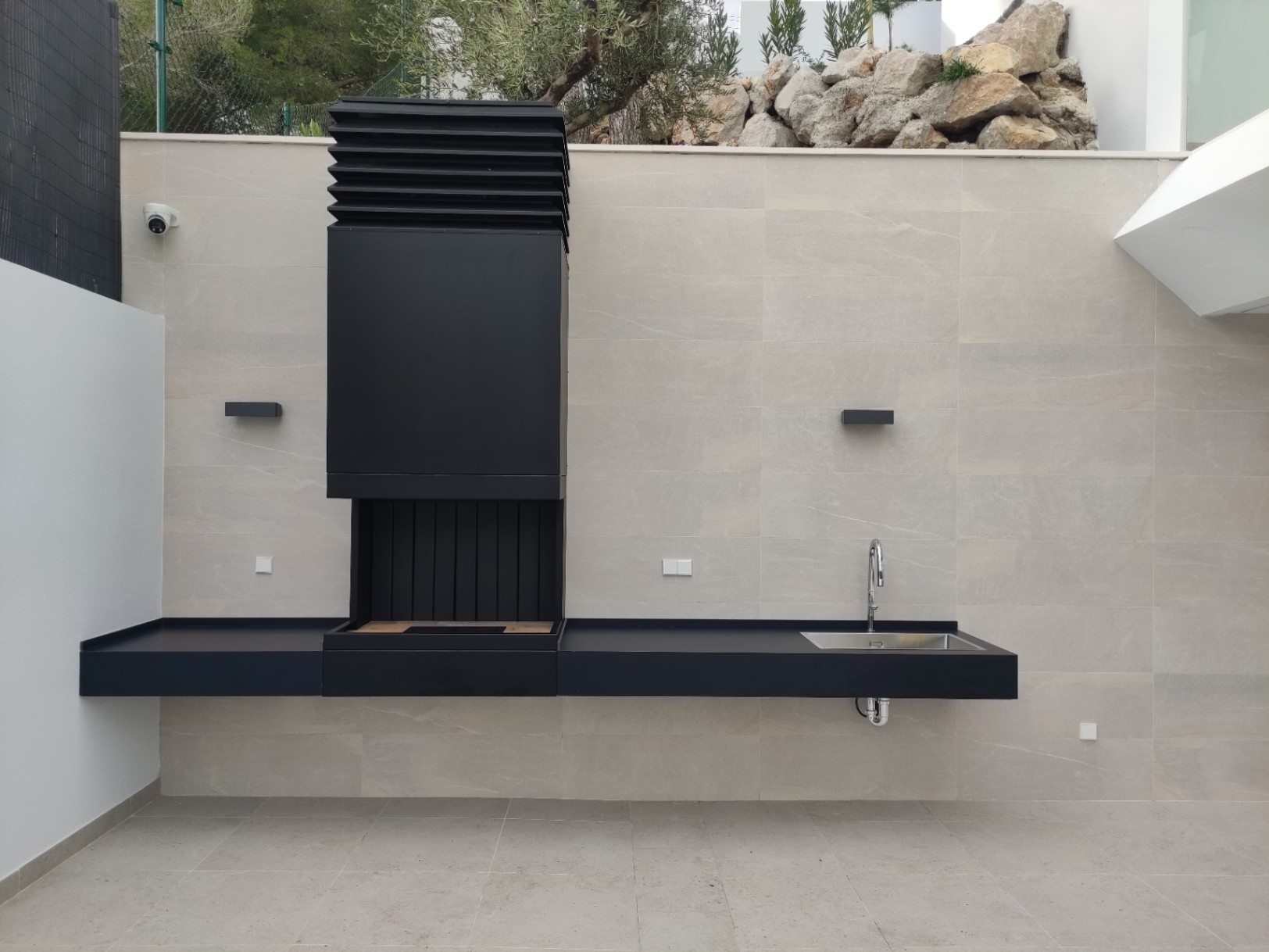 Minimalistische Außenküche mit mattschwarzem Grill, integrierter Spüle und schwebender Arbeitsplatte vor einer Wand aus großformatigen Natursteinfliesen.
