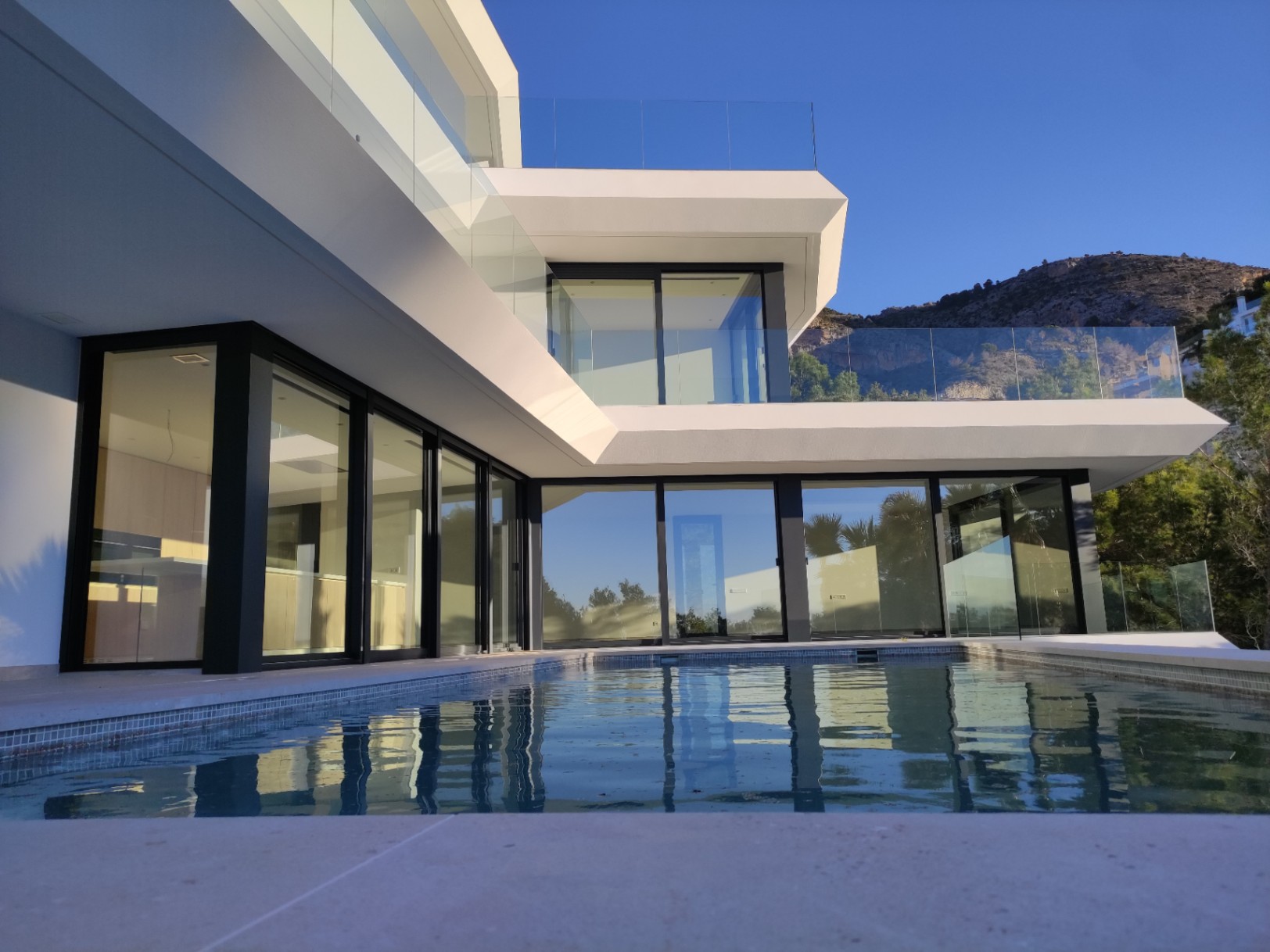 Moderne Fassade mit bodentiefen, schwarz gerahmten Fenstern und einem Infinity-Pool, der die geometrische Architektur vor der Kulisse von Altea Hills widerspiegelt.