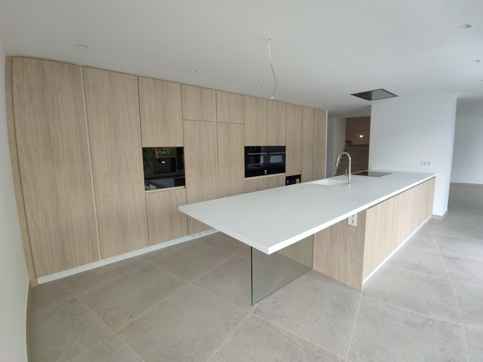 Moderne Küche mit Holzfronten, integrierten Geräten und einer weißen Kochinsel mit gläsernem Stützfuß auf großformatigen Bodenfliesen.