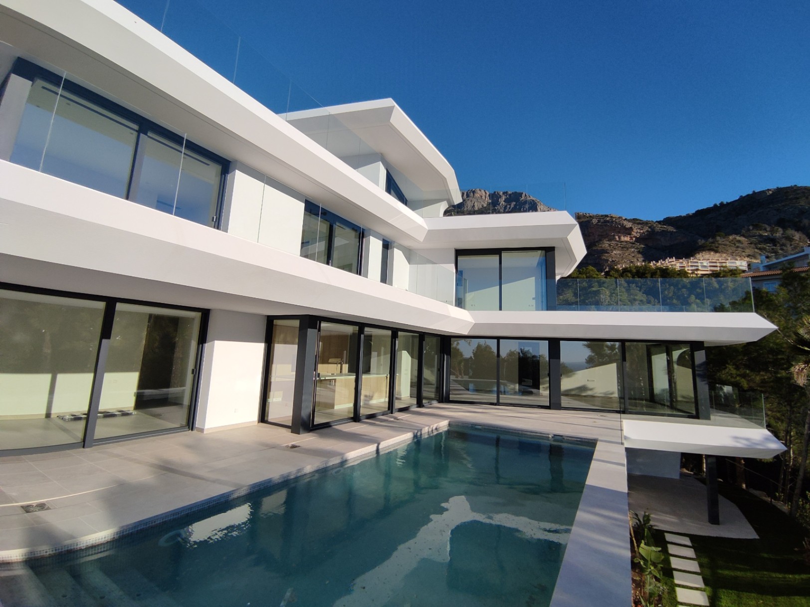Moderne weiße Fassade mit auskragenden Ebenen, bodentiefen Glasfronten und einem Infinity-Pool vor der Kulisse der Berge von Altea Hills.