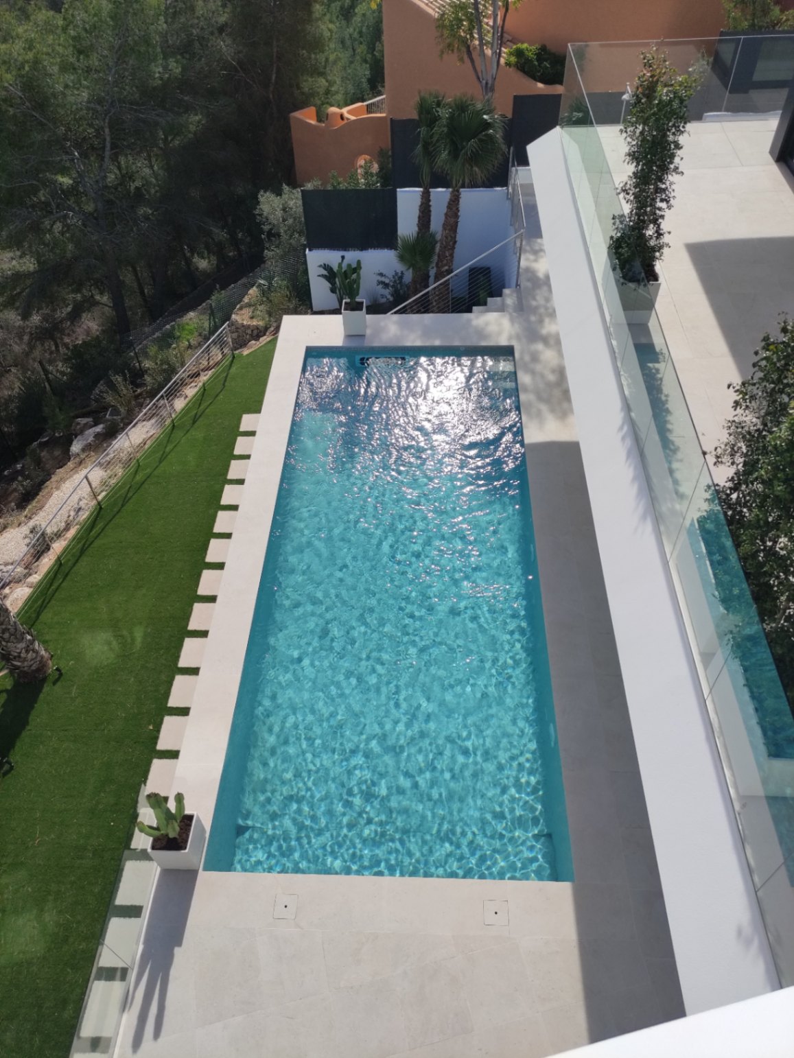 Rechteckiger Swimmingpool mit klarem blauem Wasser, umgeben von hellen Steinterrassen, Kunstrasen mit Trittsteinen und modernen Glasgeländern.