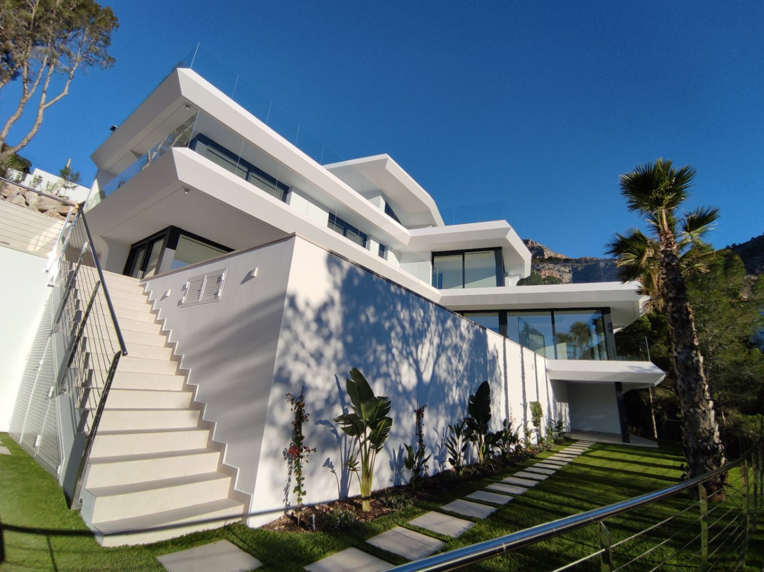 Zeitgenössische mehrstöckige Außenfassade mit weißen Linien, Glasgeländern und einem Garten mit Steinpfad und mediterranen Pflanzen.