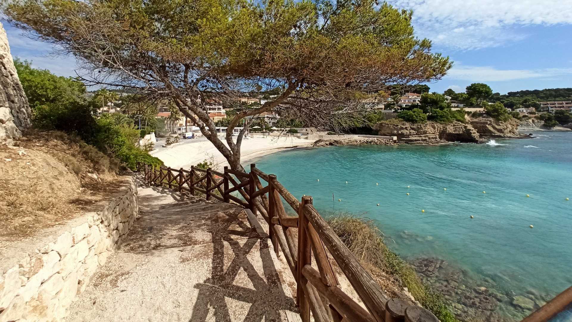 Küstenpfad mit Holzgeländer und Natursteinmauern, der direkt zum Sandstrand von La Fustera und dem türkisfarbenen Mittelmeer führt.