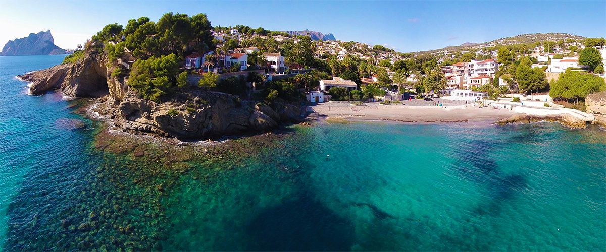 Luftaufnahme des Strandes La Fustera mit türkisblauem Wasser, Felsklippen und dem Peñón de Ifach im Hintergrund, eingebettet in mediterrane Pinien und Villen.