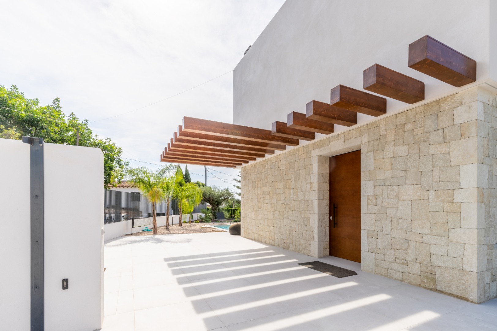 Haupteingang mit Steinmauer und Holzpergola, die Schatten auf den Boden wirft.