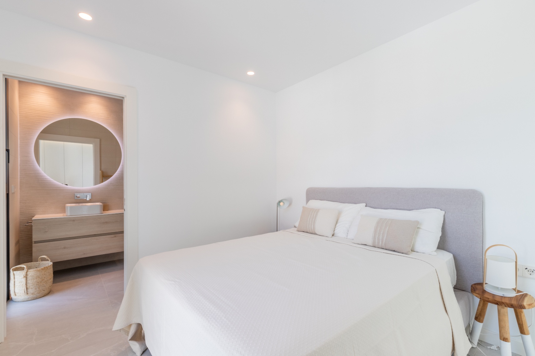 Helles Schlafzimmer mit Bad en Suite. Doppelbett mit grauem Kopfteil und weißer Bettwäsche. Badezimmer mit rundem, beleuchtetem Spiegel.