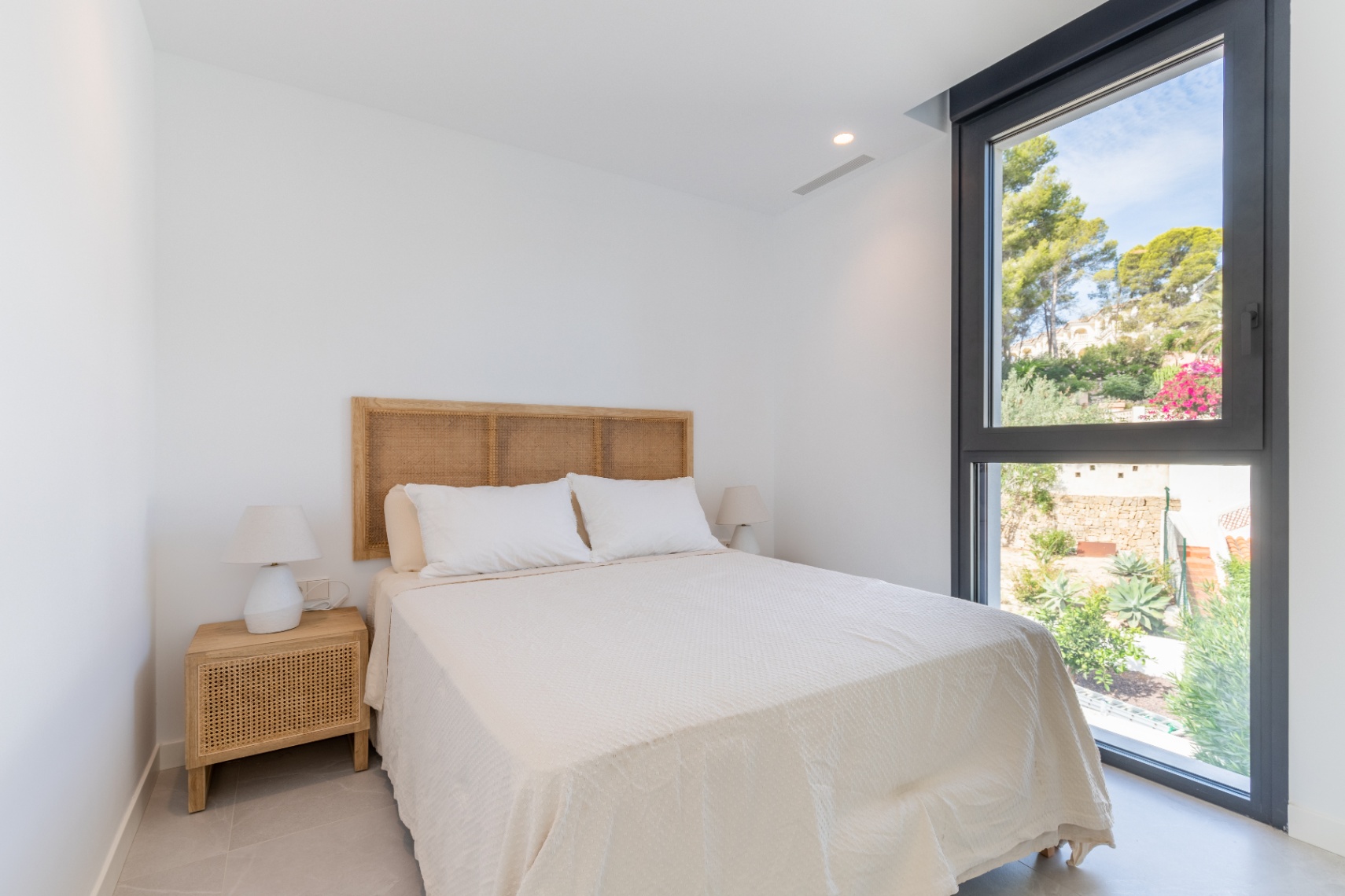 Helles Schlafzimmer mit Rattan-Kopfteil und Gartenblick aus dem schwarz gerahmten Fenster.