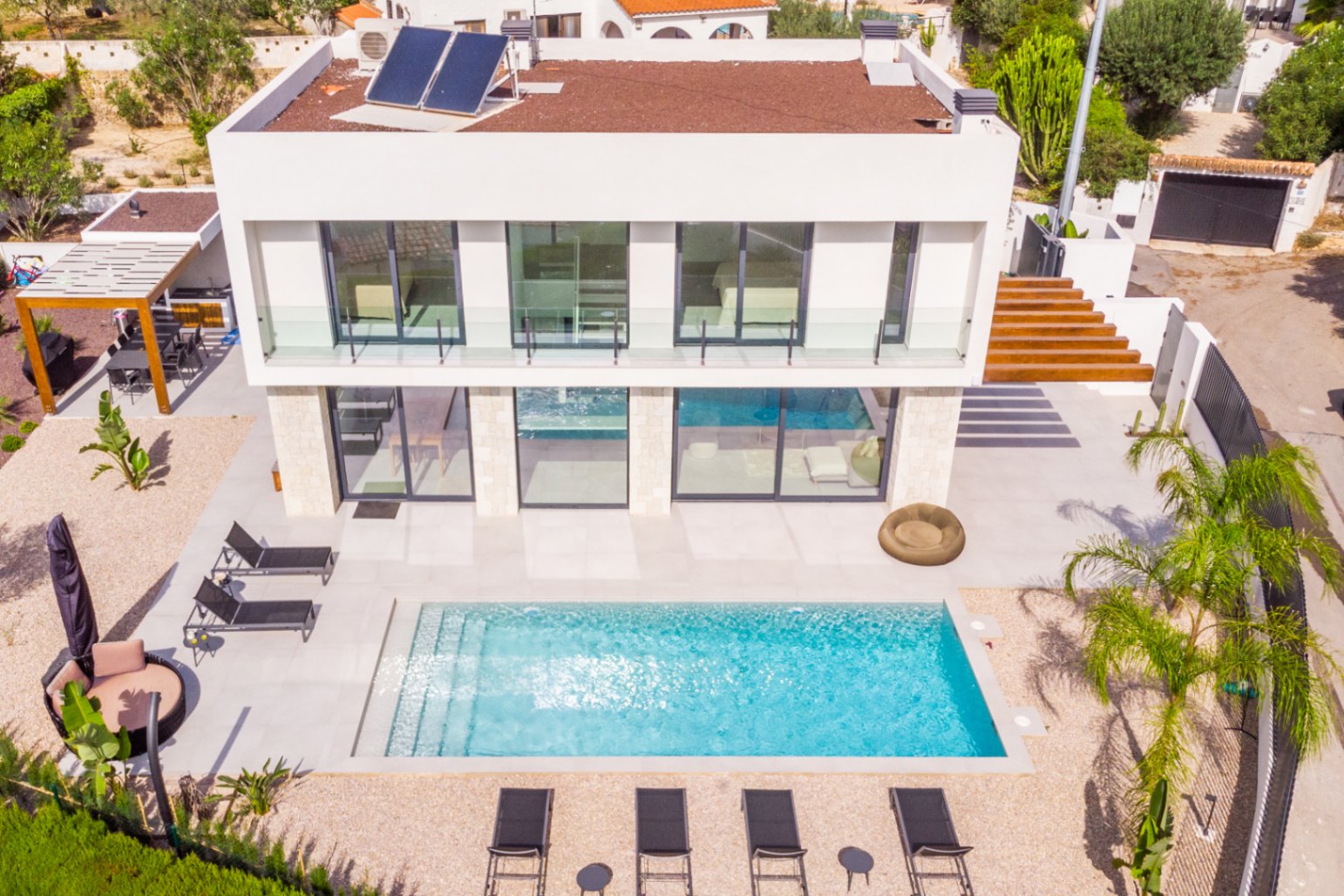 Luftaufnahme der Villa mit rechteckigem Pool, umgeben von Sonnenliegen und hellem Kies. Modernes Design mit Solarpaneelen.