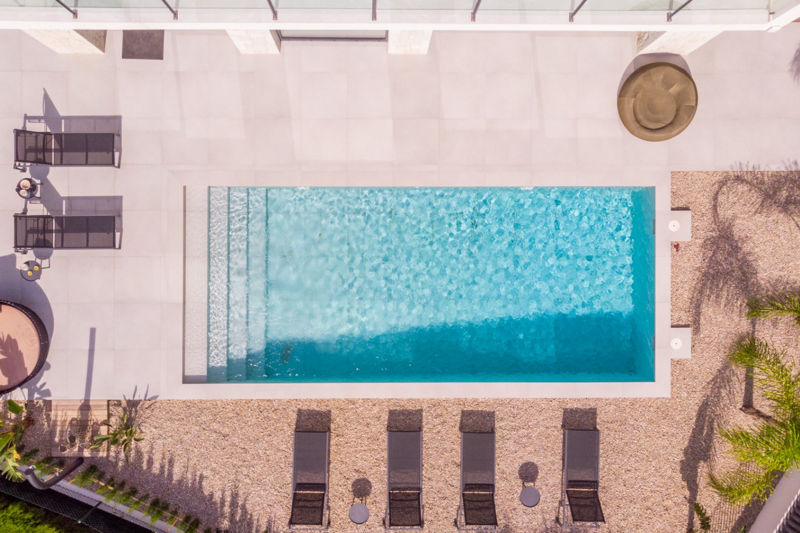 Luftaufnahme des rechteckigen Pools mit Stufen, umgeben von hellen Fliesen und Kies. Liegestühle und Vegetation vervollständigen das Design.
